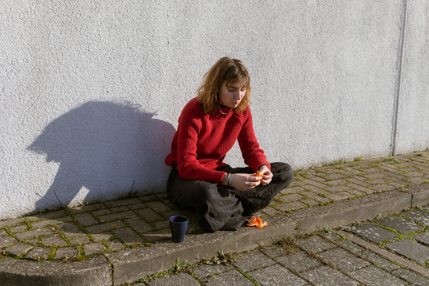 Junge Frau sitzt auf dem Bürgersteig vor einer weißen Wand, trägt roten Pullover und schwarze Hose, schält eine Orange