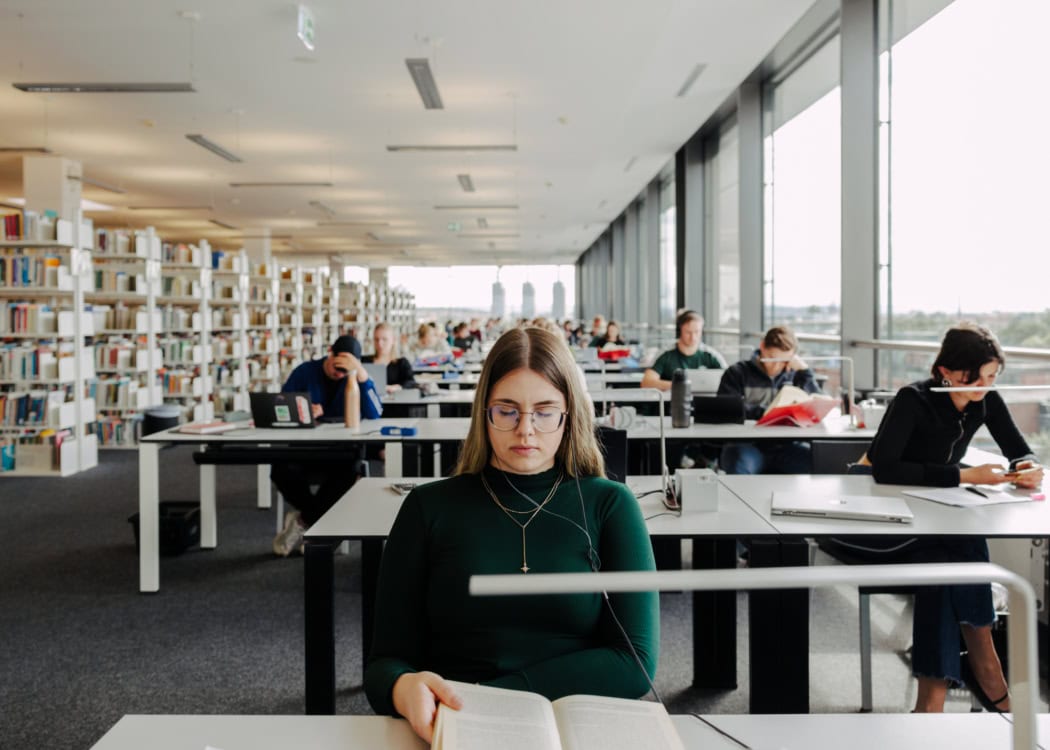 Die Studentin Livia Sommer sitzt tagsüber in der Universitätsbibliothek. In der Hand hält sie ein Buch, ihre Augen sind geschlossen.