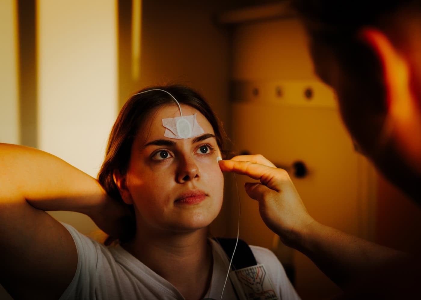 Sommer sitzt in ihrem Krankenhauszimmer. Nachdem die Haut entfettet wurde, bringt die Krankenpflegerin Marie Drebing die ersten zwei Elektroden im Augenbereich von Sommer an. Sie sorgen dafür, dass die Augenbewegungen getrackt werden können. Das Licht scheint ihr warm ins Gesicht.