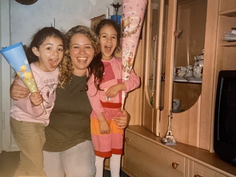 Auf dem Foto sieht man eine Frau, die links und rechts ihre Töchter im Arm hält. Beide Kinder haben braune Haare und lächeln wie ihre Mutter sehr glücklich in die Kamera. Die Tochter auf der linken Seite hält eine kleine blaue Schultüte in der Hand, die Tochter auf der rechten Seite eine größere, rosa Schultüte.