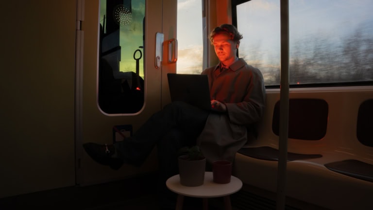 Ein junger Mann mit hellbrauen Locken sitzt in der Straßenbahn. Sein Rücken zeigt zum Fenster. Er hat seine Beine überschlagen und auf seinem Schoß befindet sich ein Laptop. Der Bildschirm des Laptops lässt sein Gesicht in orangenen Licht leuchten. Neben ihm steht ein kleiner weißer Beistelltisch, auf dem eine Pflanze und eine Tasse abgestellt sind. In seinem Ohr trägt er Bluetooth-Kopfhörer. Er ist am arbeiten und schaut auf seinen Laptop. Draußen wird es langsam dunkel.
