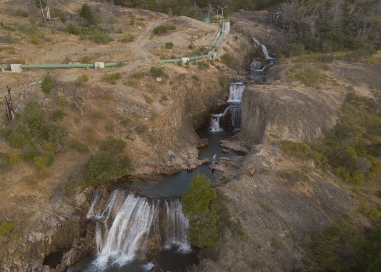 Ein Bild des Los Maquis Flusses im chilenischen Puerto Guadal mit malerischen Wasserfällen und einem grünen Rohr, das für ein neu gebautes Wasserkraftwerk installiert wurde. Die Wasserfälle stürzen in Kaskaden über Felsen hinab, umgeben von ausgetrockneter Vegetation. Das grüne Rohr ist gut sichtbar und schlängelt sich durch die Landschaft, um Wasser zum Kraftwerk zu leiten.