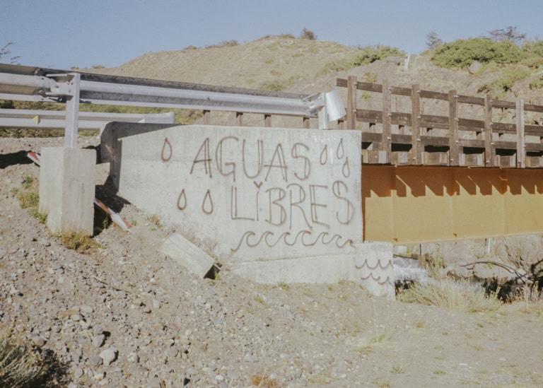 Ein Graffiti auf einer Betonwand unter einer Brücke zeigt den Text "AGUAS LIBRES" (freie Wasser). Um den Text herum sind mehrere Tropfen und Wellenlinien gemalt. Die Umgebung ist eine trockene, steinige Landschaft mit einigen Sträuchern und einem Hügel im Hintergrund. Die Brücke selbst besteht aus Holz und Metallteilen.