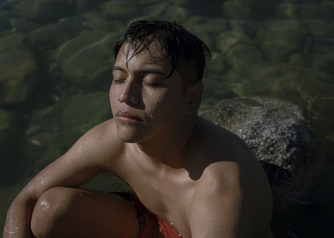 Ein junger Mann sitzt mit geschlossenen Augen in einem klaren, flachen Wasser, das von Felsen umgeben ist. Er trägt rote Badebekleidung und hat nasses Haar, während Sonnenlicht auf sein Gesicht scheint, wodurch er einen entspannten und friedlichen Ausdruck hat.